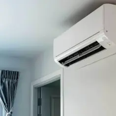 Biała jednostka klimatyzacyjna zamontowana na jasnej ścianie, obok drzwi i okna z zasłoną.