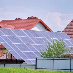 Duży panel fotowoltaika na gruncie przed domem z czerwonym dachem. Zielona trawa i płot.